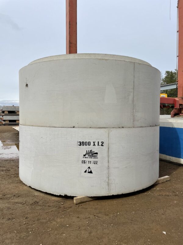 Large concrete cylinder with text "3000 X 1.2" and "Miller Precast." Industrial background, steel beams visible, outdoors on muddy ground.