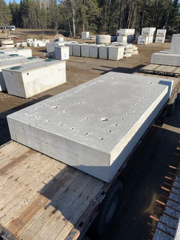 Concrete blocks and septic tanks are arranged outdoors. A large block is on a truck in a wooded area with clear skies.