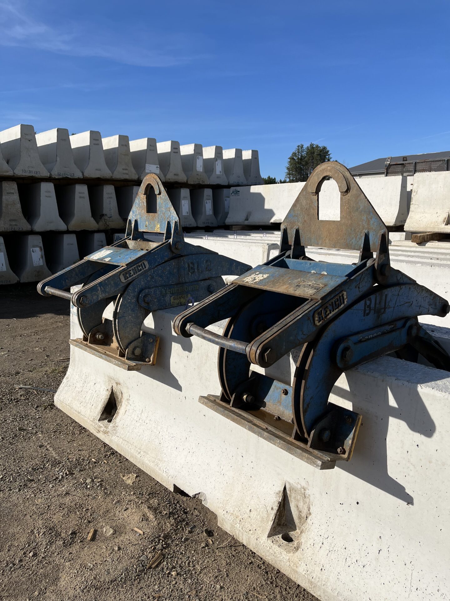 Concrete barriers with metal clamps are stacked on a construction site, under a clear blue sky. Trees and structures are visible in background.