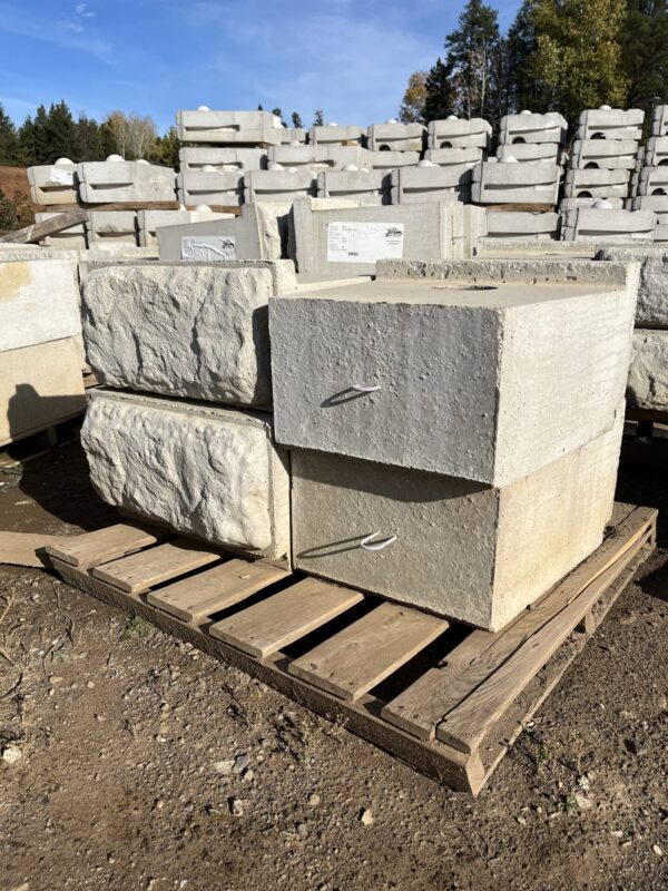 Concrete blocks stacked on wooden pallets, outdoors. Surrounding area includes trees and clear blue sky. No people, landmarks, or historical buildings visible.