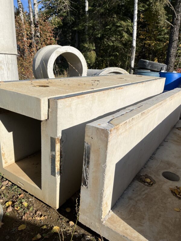 Concrete construction materials, including rectangular slabs and circular pipes, lie on the ground surrounded by trees and scattered leaves under a clear blue sky.