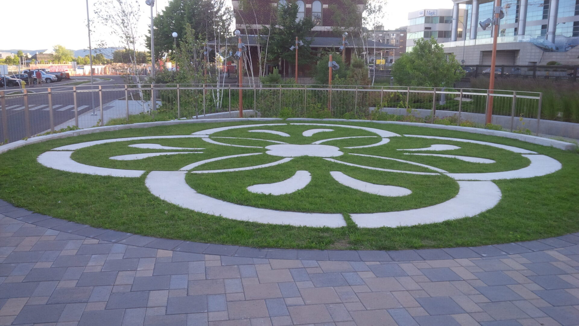 Ornate patterned garden with circular grassy design, surrounded by pavement. Nearby, a brick building and modern glass structure are visible. Quiet urban setting.