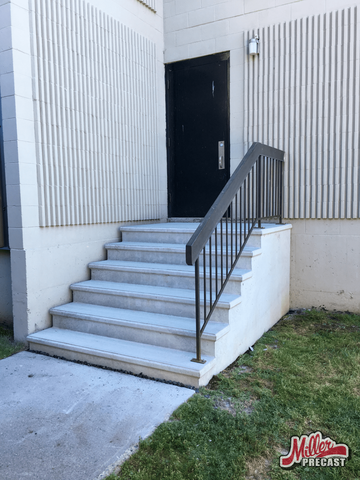 Concrete steps with a black metal railing lead to a dark door on a beige building. No people, landmarks, or historical buildings visible.