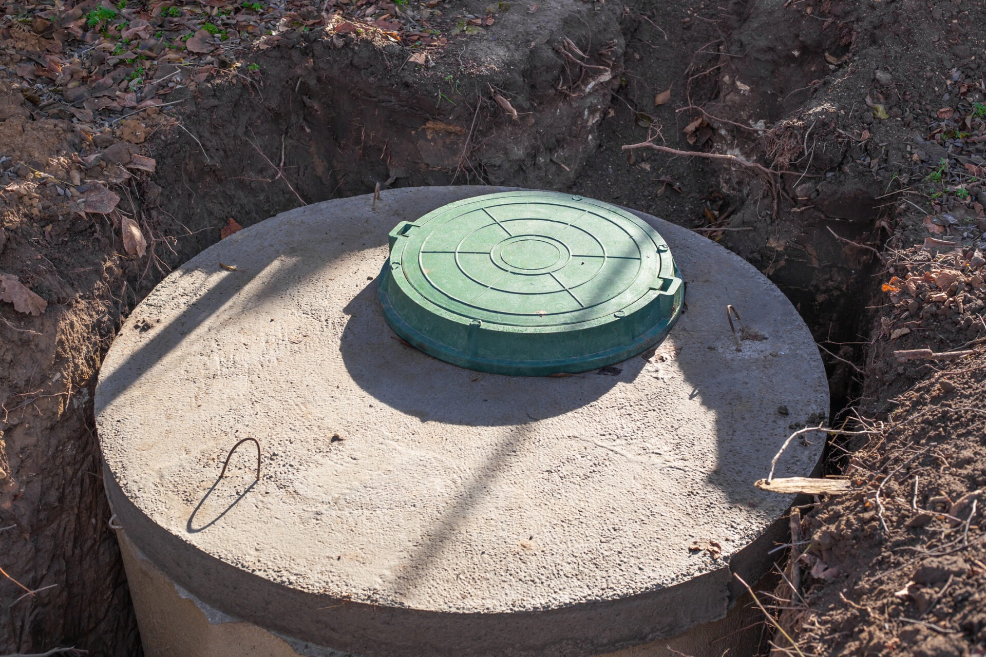 Concrete septic tank with a green plastic cover partially buried in soil, surrounded by leaves and dirt, under bright sunlight.