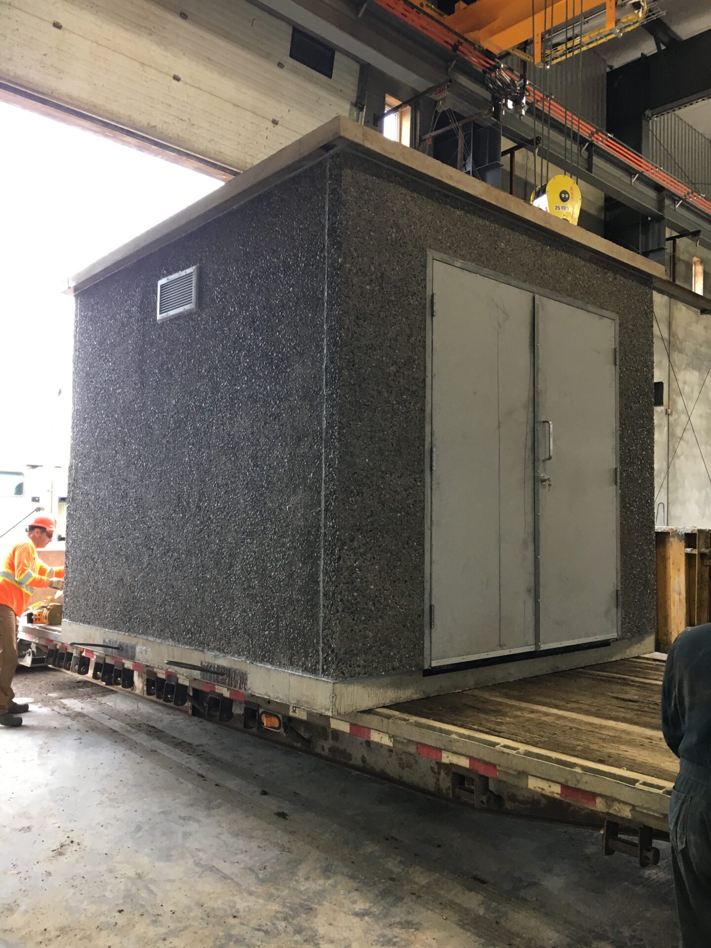 A concrete structure is loaded onto a flatbed truck inside an industrial warehouse. Two people in safety gear are assisting.