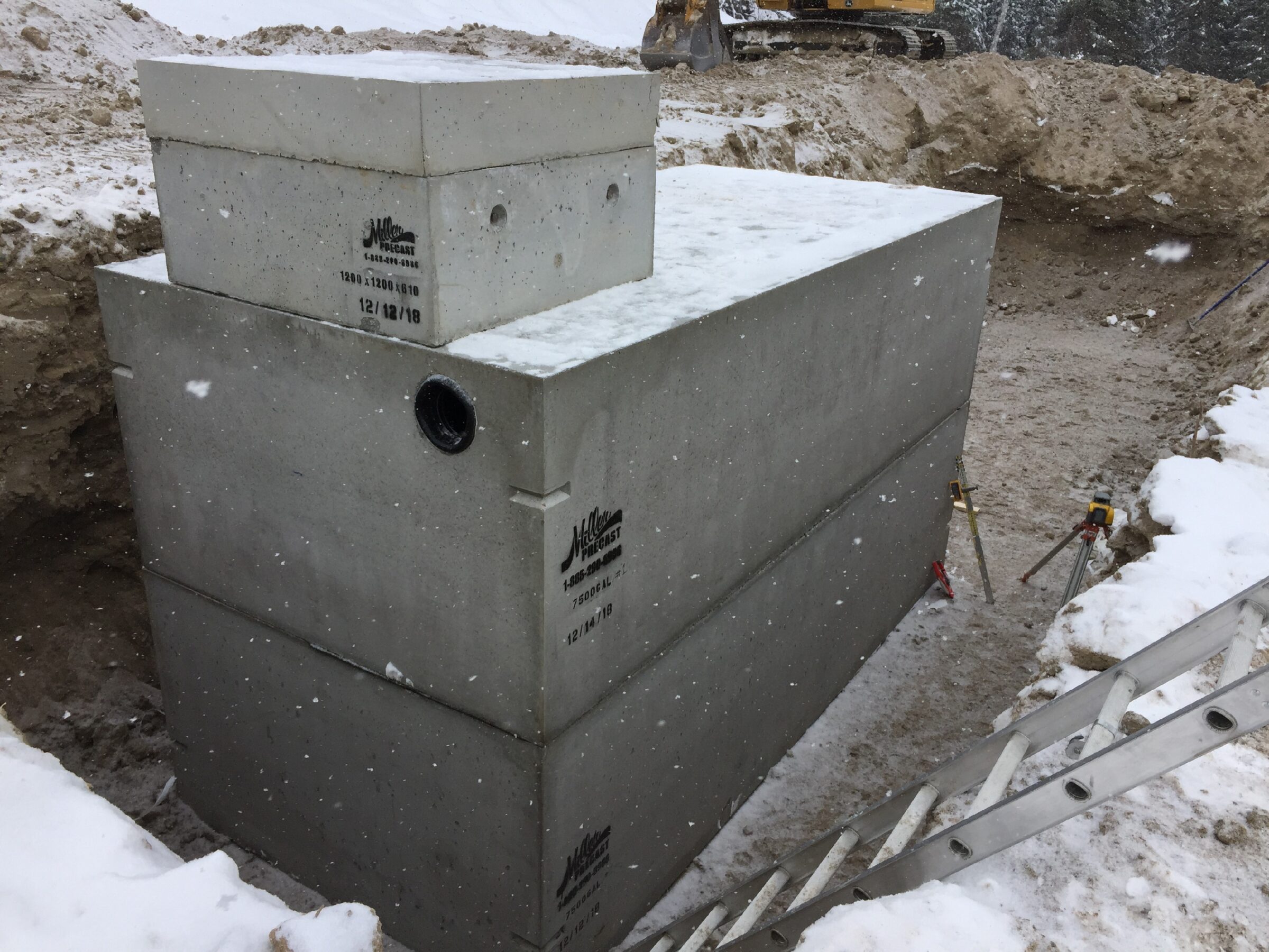 A large concrete septic tank is installed in a snowy excavation site, with a ladder and construction equipment nearby.