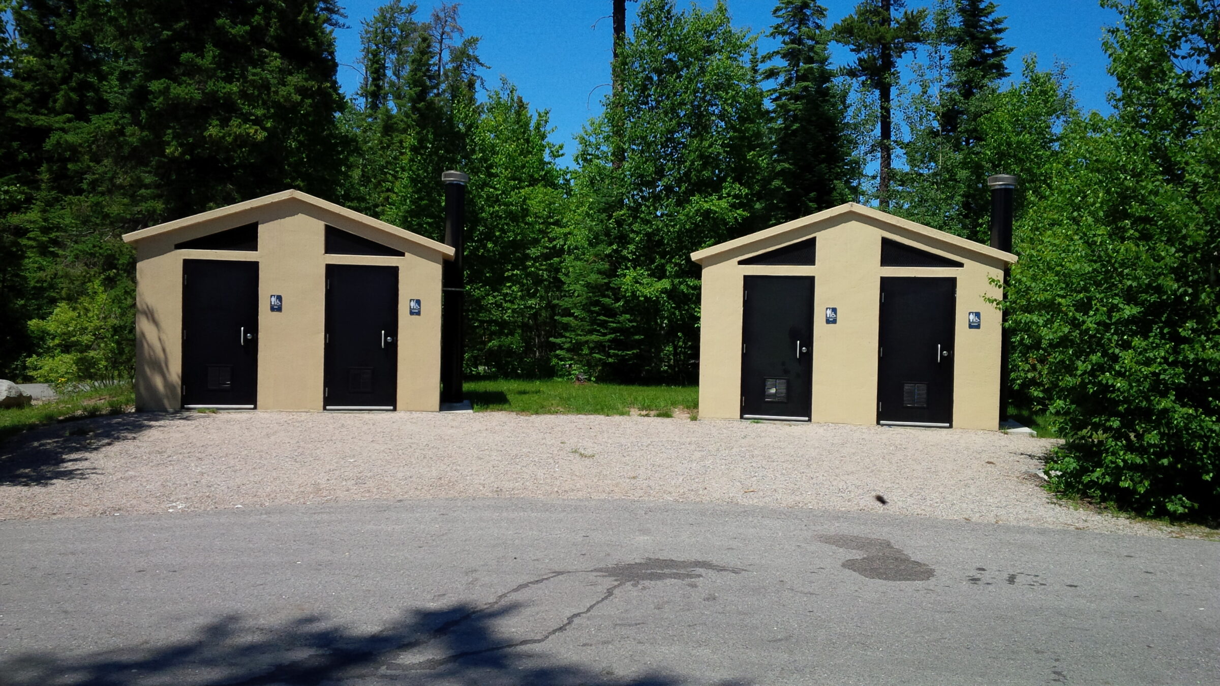 Two tan restroom buildings with black doors in a forested area, surrounded by trees, under a blue sky on a sunny day.