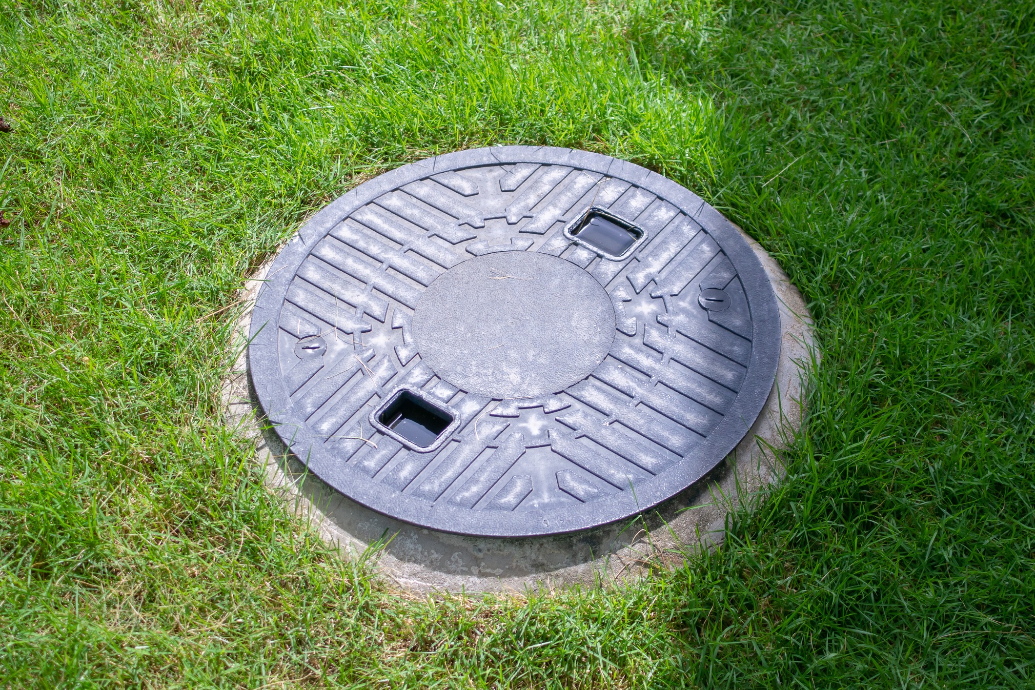 Metal manhole cover embedded in a grassy lawn, featuring geometric patterns and two rectangular openings, surrounded by lush, green grass.