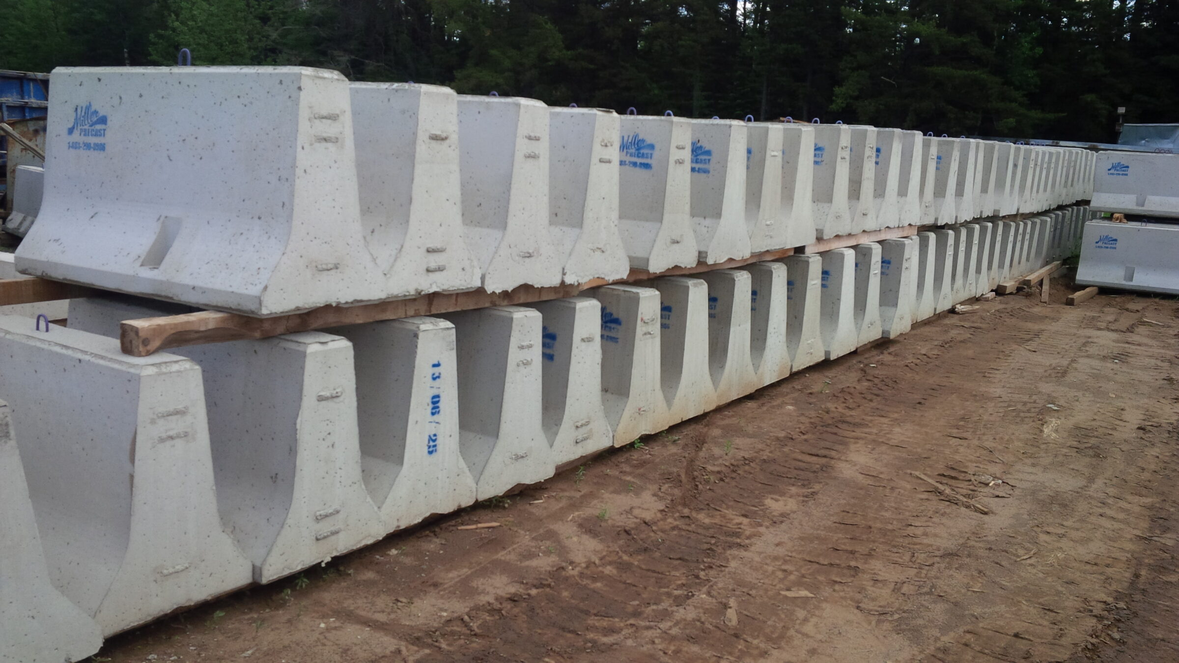 Rows of stacked concrete barriers on dirt ground, surrounded by trees, with blue logos and contact information visible on each barrier.