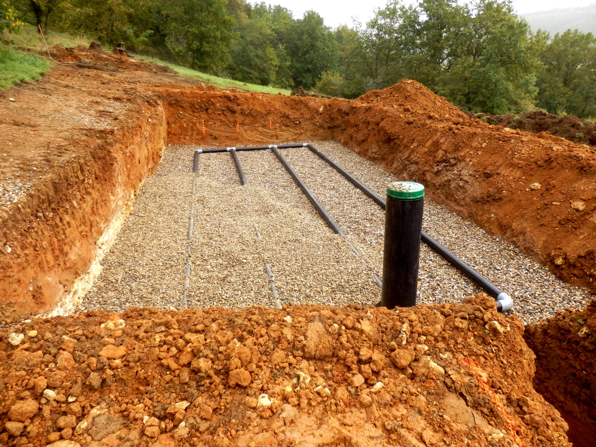 A freshly excavated septic system trench with pipes installed, surrounded by red earth. Forested area visible in the background. No people present.