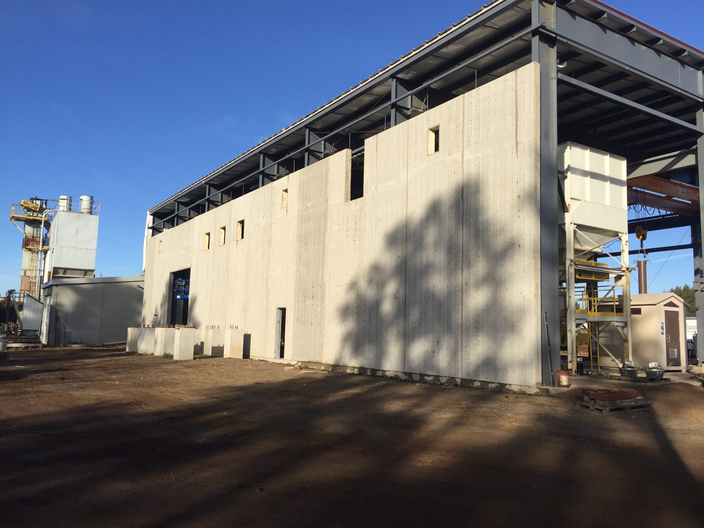 Industrial building under construction with exposed steel framework and concrete walls. Clear blue sky and nearby machinery visible in the background.