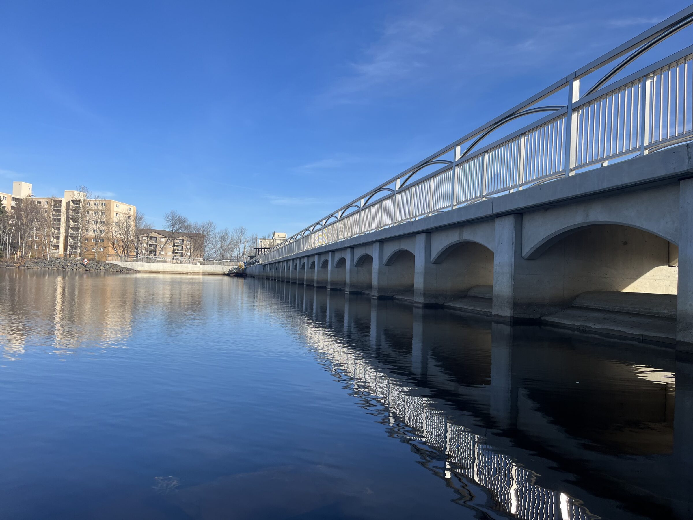 A modern bridge spans a calm river, with apartment buildings in the background under a clear blue sky. Reflection details enhance the scene.