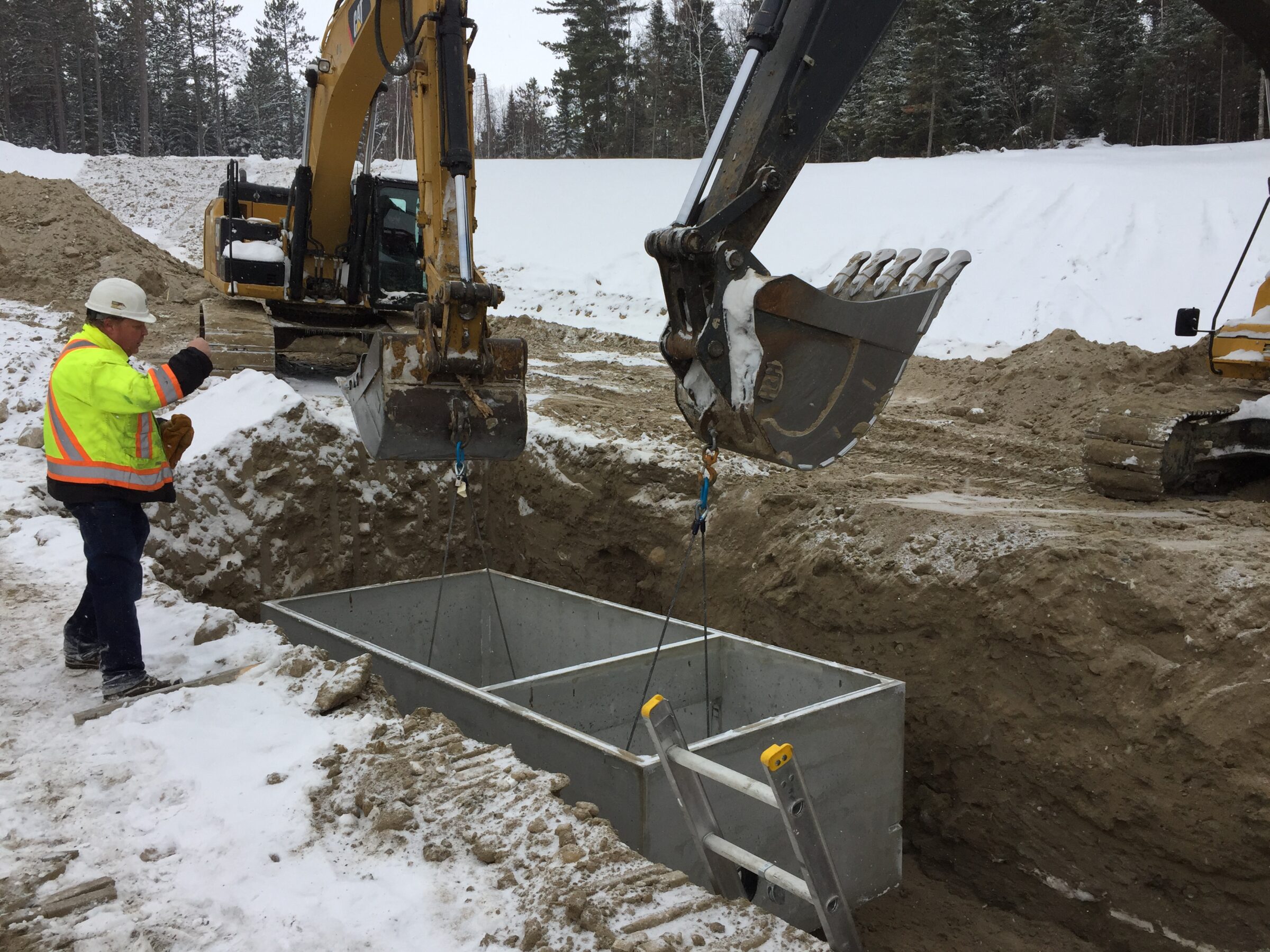 A person in a safety jacket observes an excavator lowering equipment into a snowy construction site surrounded by trees.