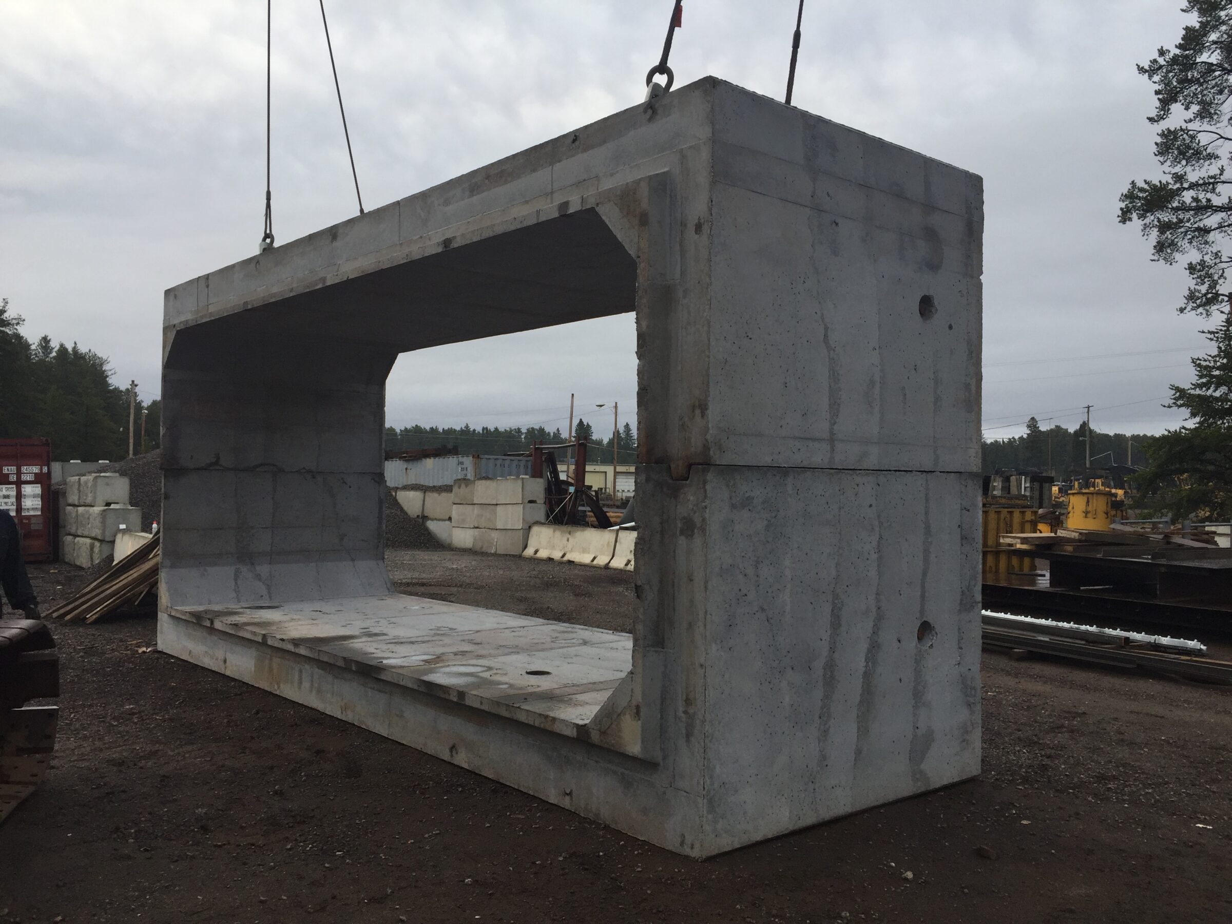 Large concrete structure being lifted by crane in industrial area with shipping containers and construction materials in the background, surrounded by trees.