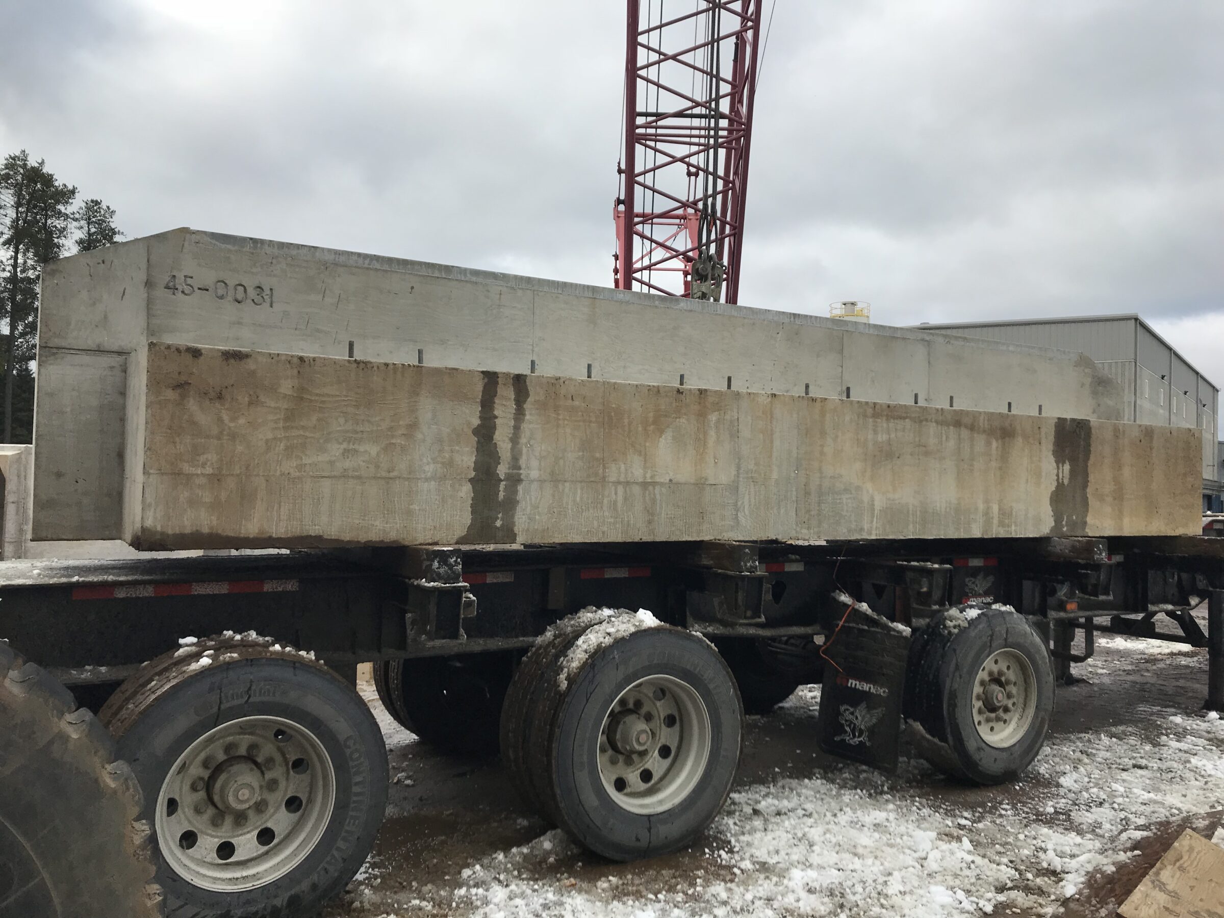 A large concrete block sits on a flatbed truck in a snowy construction site, with a crane visible in the background.