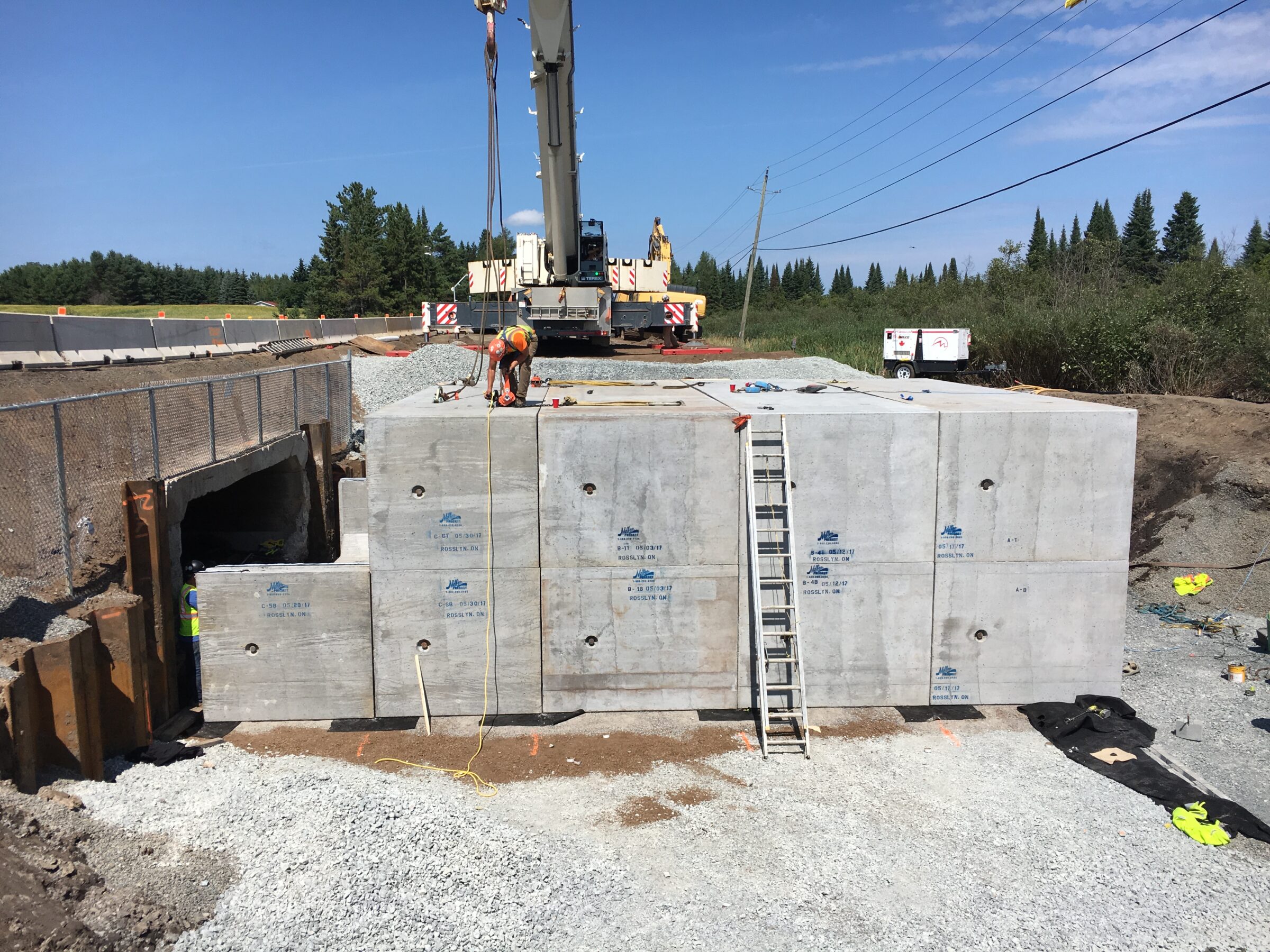 A person works on a large concrete structure at a construction site, surrounded by trees and equipment, under a clear blue sky.