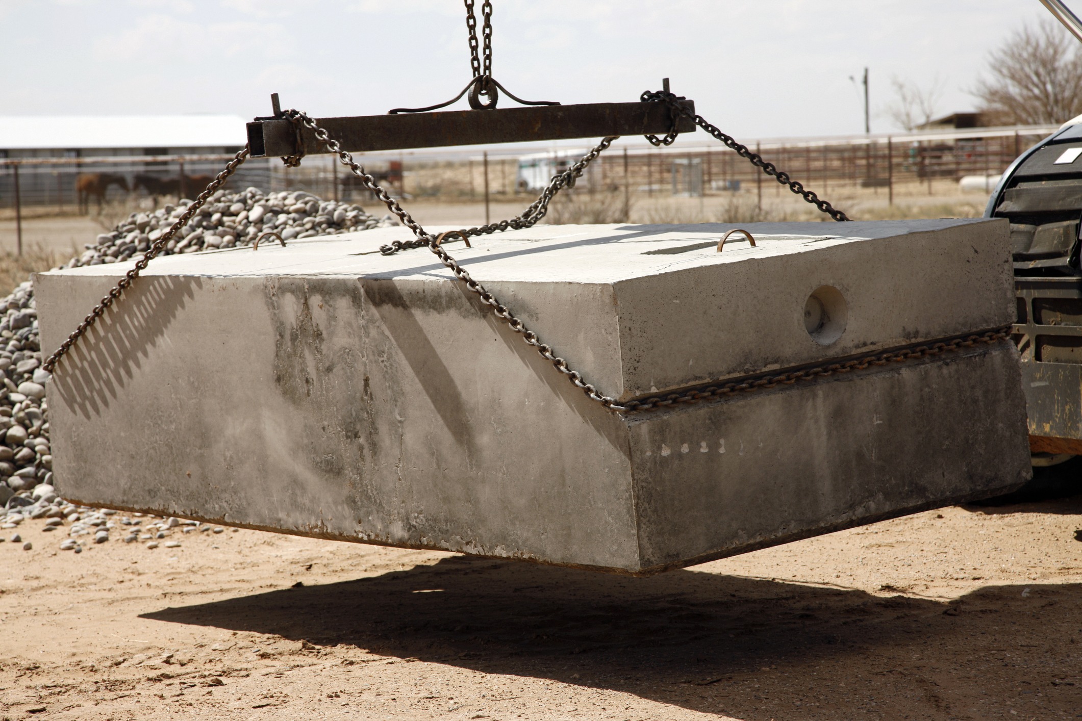 A large concrete block is suspended by chains, lifted by machinery in a dusty outdoor area, with a horse in the background.
