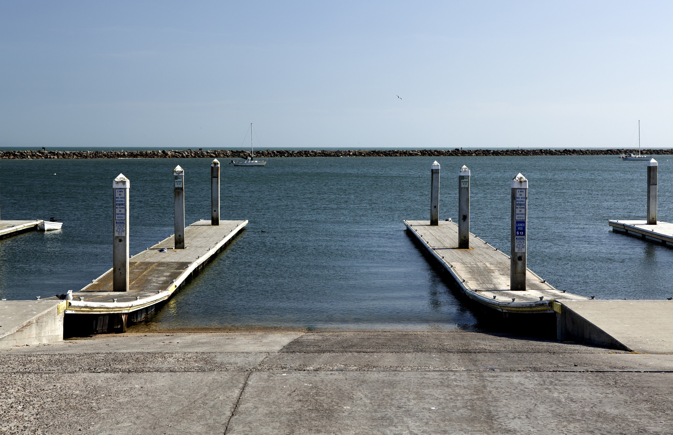 Two empty docks extend into calm water, with a sailboat and distant rock barrier under a clear sky. Tranquil, open seascape scene.