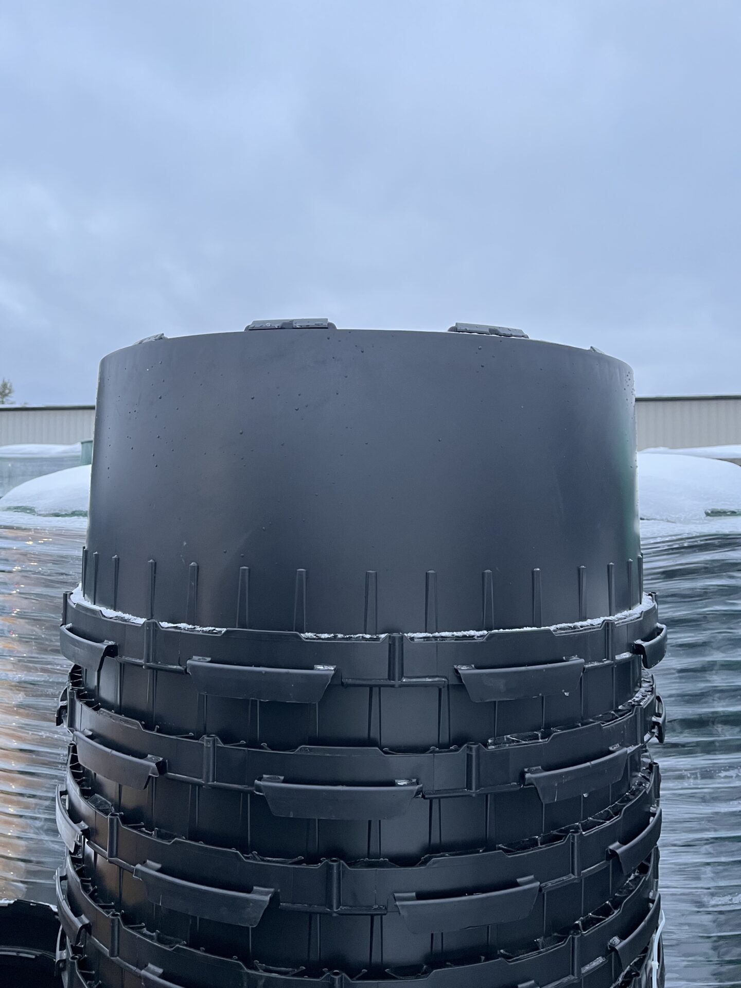 A stack of large, black plastic containers under an overcast sky, with part of a building visible in the background.