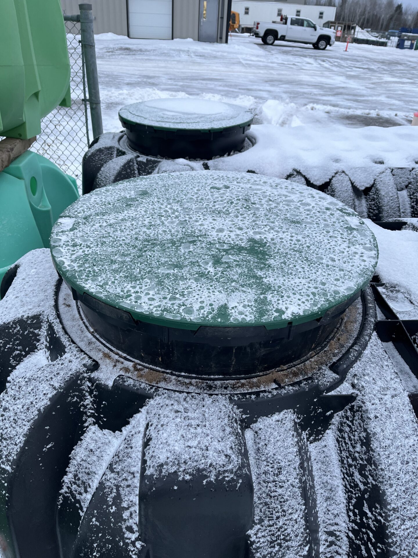 Snow-covered plastic containers and equipment outdoors near a fenced area. Buildings and a white truck are visible in the snowy background.