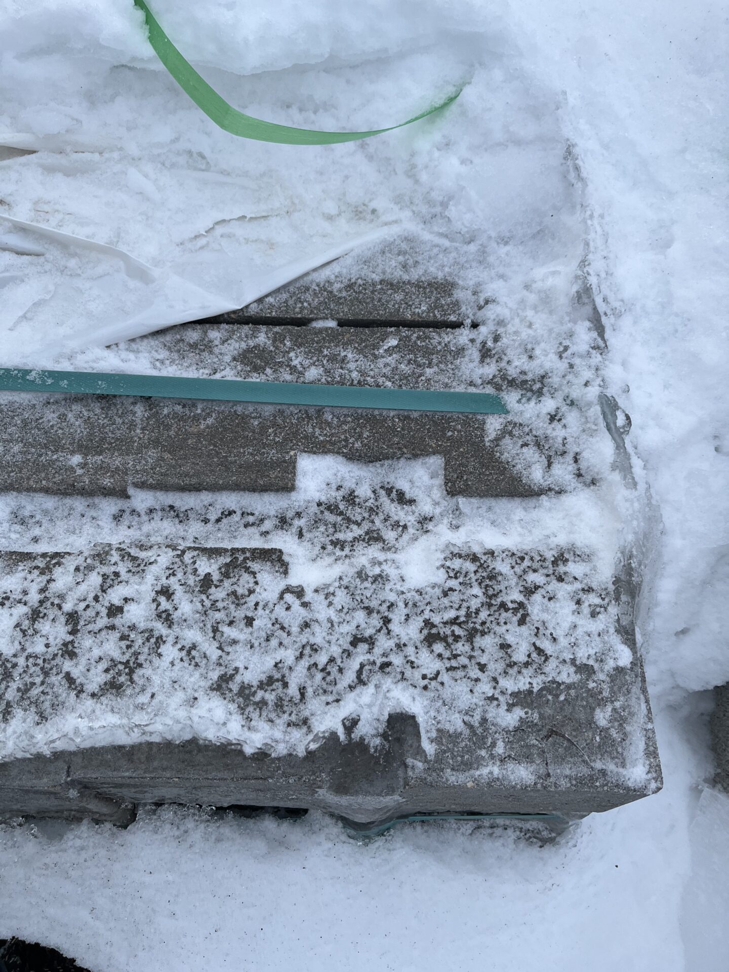 Concrete blocks partially covered in snow and ice, with green and white plastic straps lying on top in an outdoor setting.