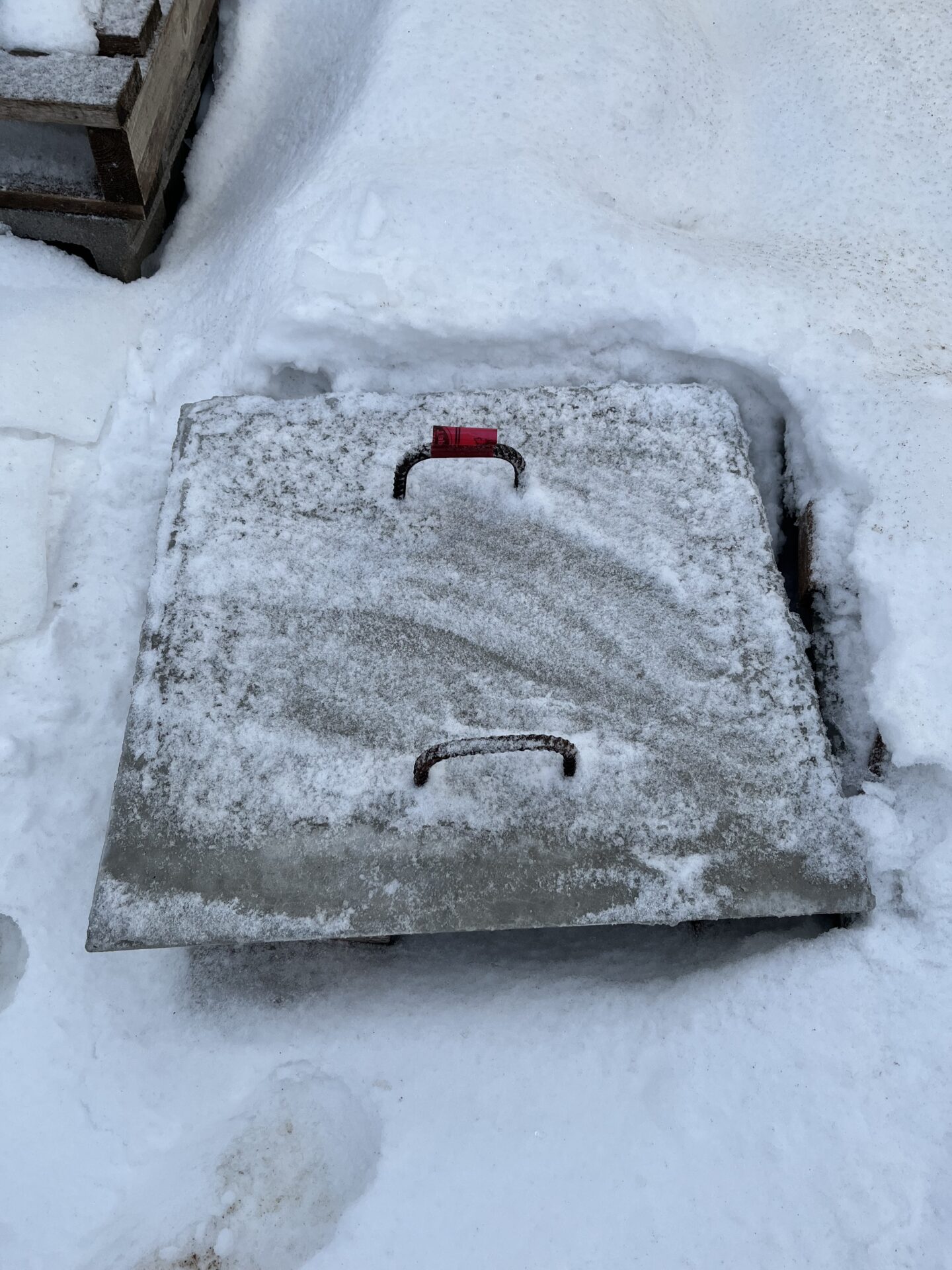 A snow-covered metal hatch with a red lock is embedded in the ground, next to wooden steps. Snow footprints are visible.