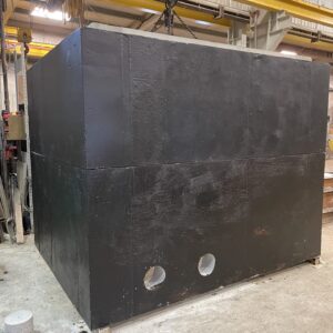 A large black industrial box with circular cutouts hangs from a yellow crane inside a warehouse with concrete floors and metal beams.