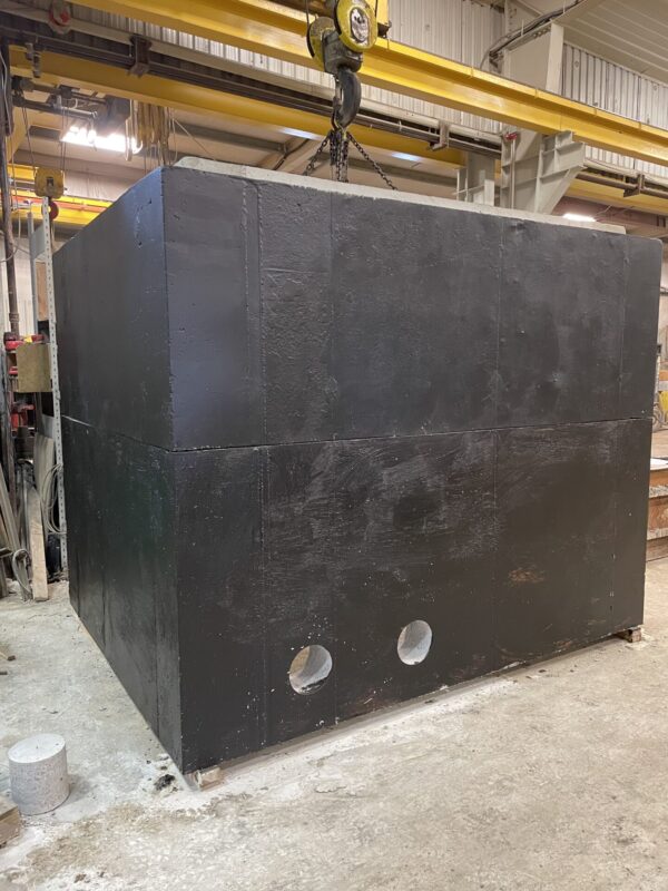 A large black industrial box with circular cutouts hangs from a yellow crane inside a warehouse with concrete floors and metal beams.