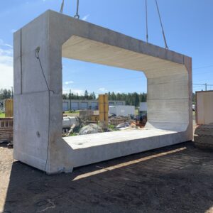 A large concrete structure is being lifted by a crane at a construction site under a clear blue sky. Industrial materials surround.