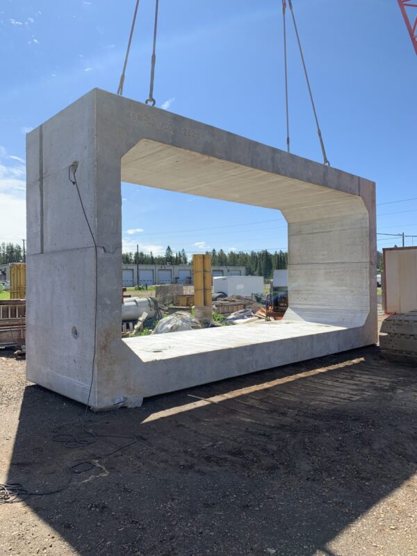 A large concrete structure is being lifted by a crane at a construction site under a clear blue sky. Industrial materials surround.