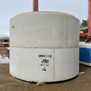 Large concrete cylinder with text "3000 X 1.2" and "Miller Precast." Industrial background, steel beams visible, outdoors on muddy ground.