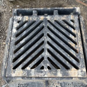 A metal drainage grate on a dirt surface, marked with "2022" and "CIP," showing wear and tear. Diagonal slots are visible.