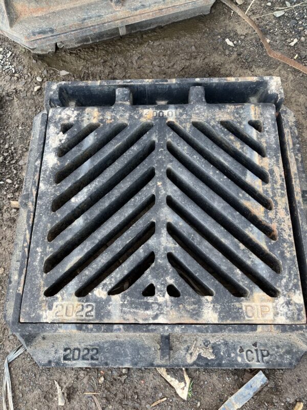 A metal drainage grate on a dirt surface, marked with "2022" and "CIP," showing wear and tear. Diagonal slots are visible.