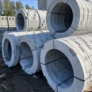 Concrete culverts stacked on dirt ground, secured with straps, in an open area surrounded by trees. No people or landmarks visible.