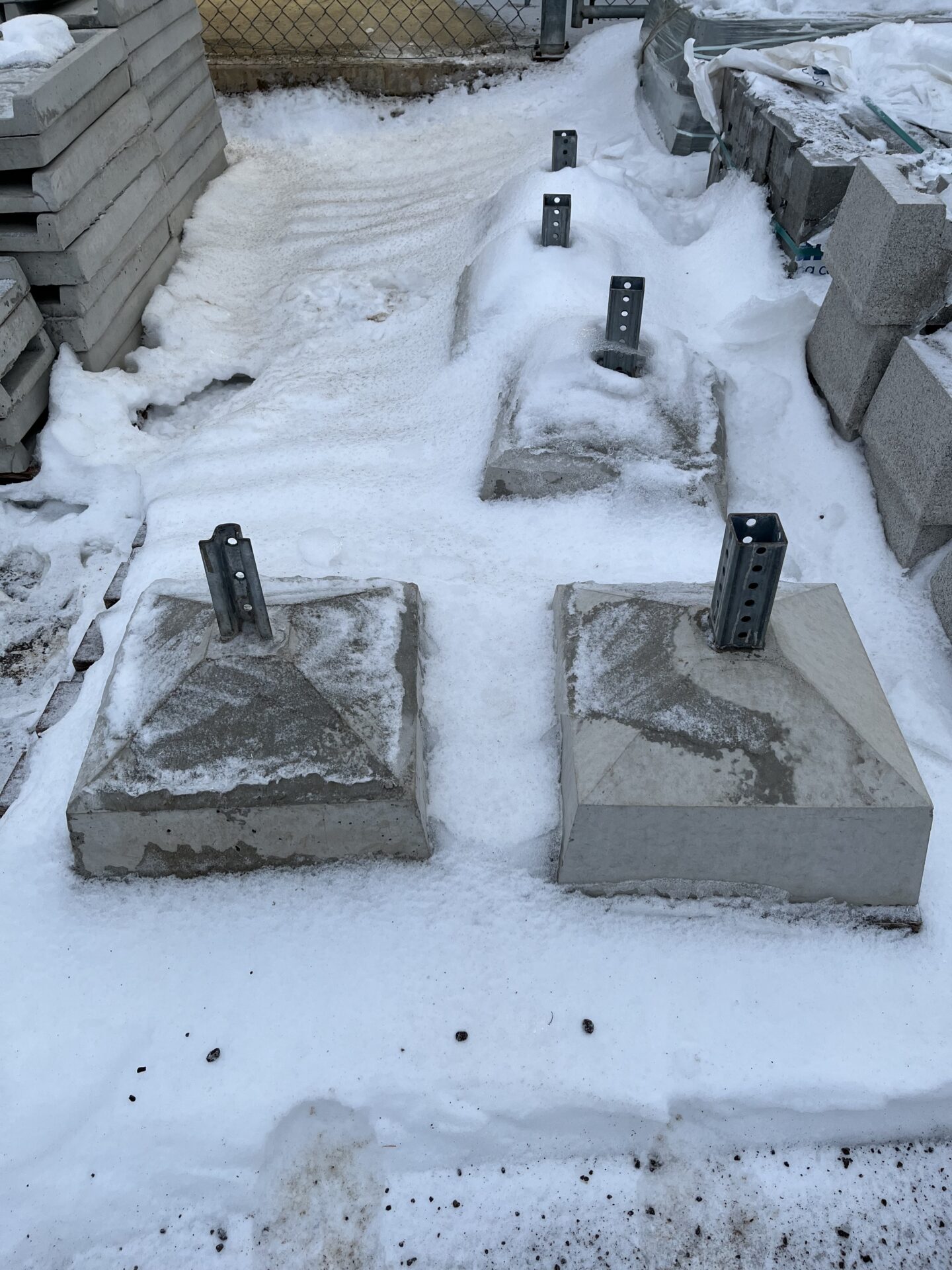 Concrete blocks with metal brackets are partially covered in snow, surrounded by stacked materials, within a construction site enclosed by a chain-link fence.