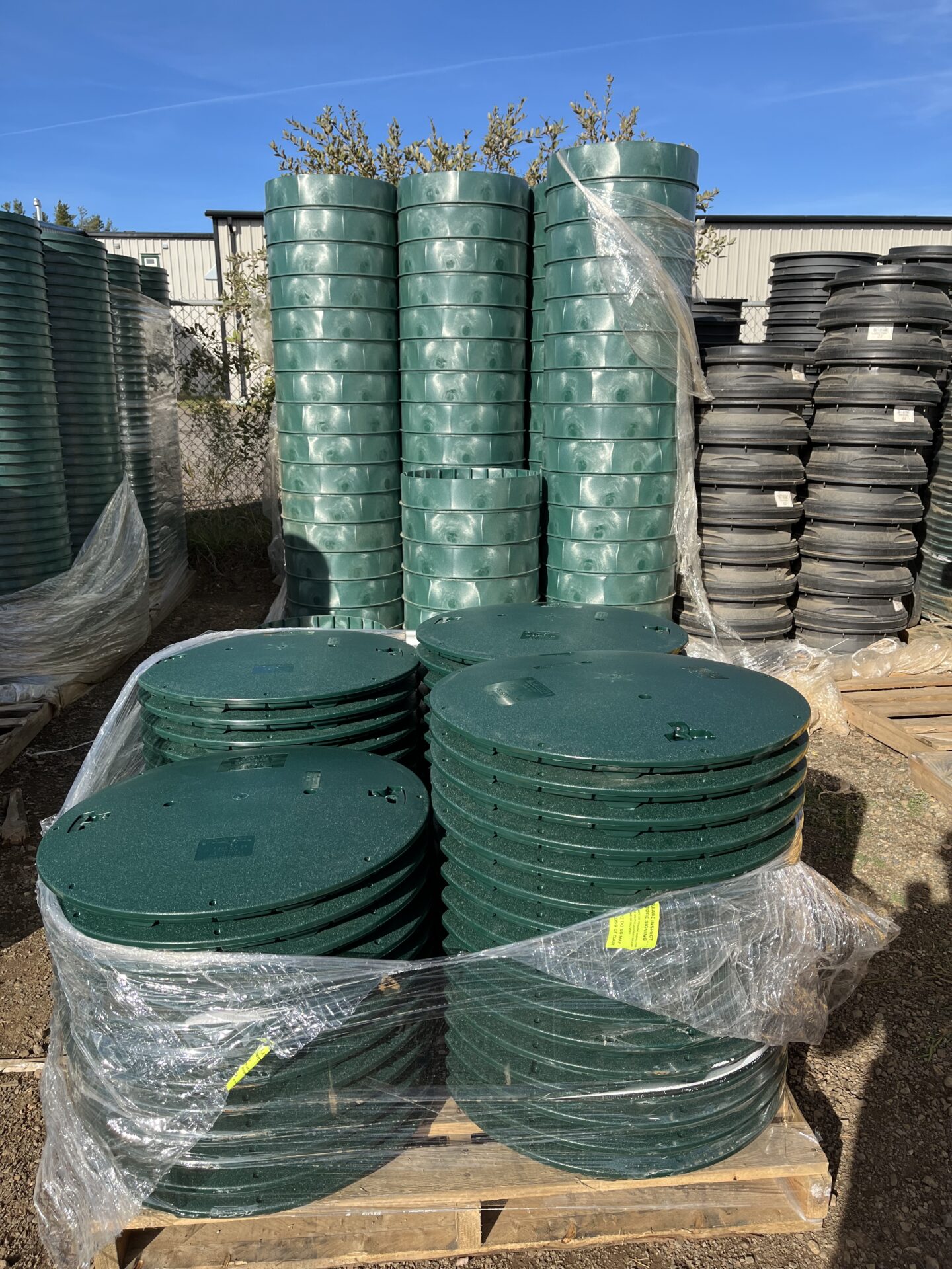 Stacks of green and black plastic lids on wooden pallets, wrapped in plastic, outside with a metal building and fence nearby under blue sky.