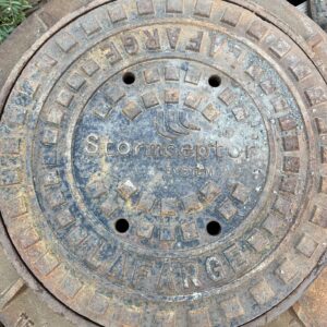 Rusty manhole cover featuring "Stormceptor System" and "Lafarge," surrounded by a wooden pallet and overgrown plants. Weathered and industrial appearance.